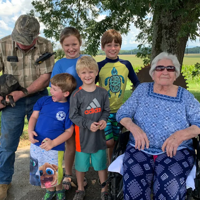 A man in a cap holds a small dog while four children pose with an elderly woman in a wheelchair under a tree on a sunny day outdoors.